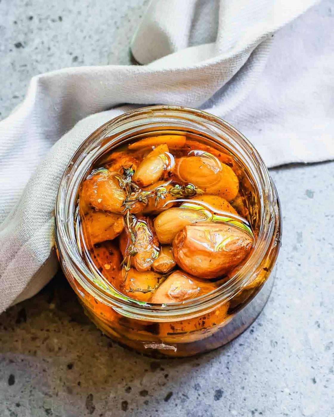 A jar filled with garlic confit marinated in oil and herbs, placed on a gray surface with a white cloth beside it.