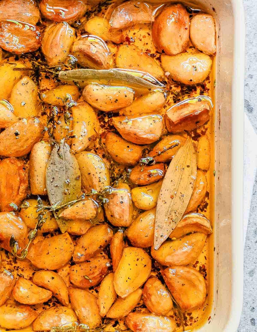 A baking dish filled with roasted garlic cloves seasoned with olive oil, bay leaves, and pepper.