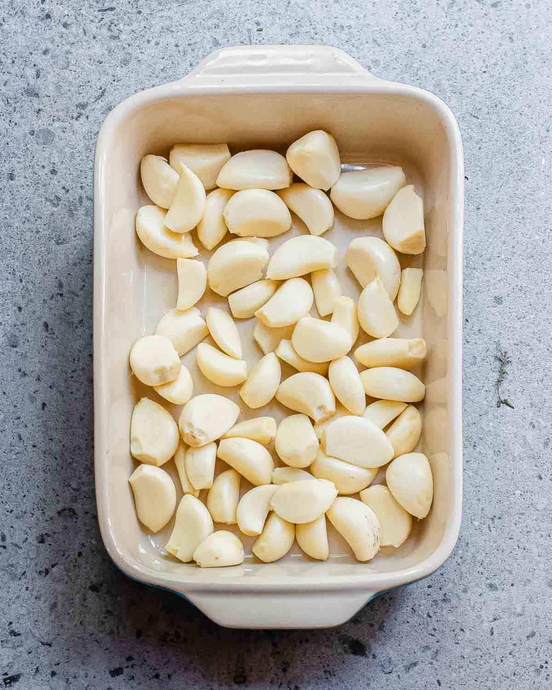 A beige rectangular dish filled with peeled garlic cloves is placed on a speckled gray surface.