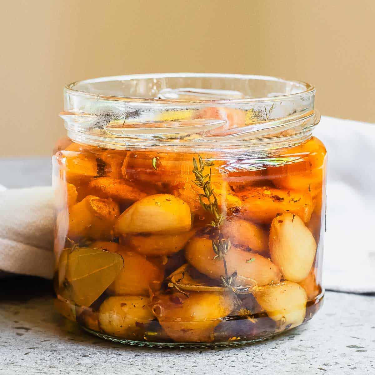 A glass jar filled with baked garlic confit and herbs in oil, placed on a gray surface.