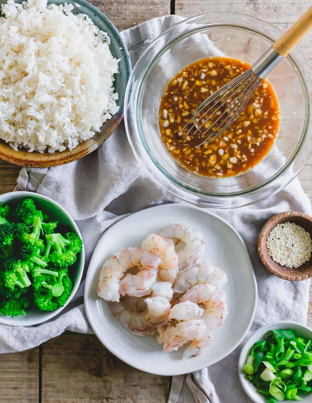 Ingredients to make General Tso's shrimp: raw deveined and peeled shrimp on a plate, General Tso's sauce with a whisk and broccoli, rice and sesame seeds.
