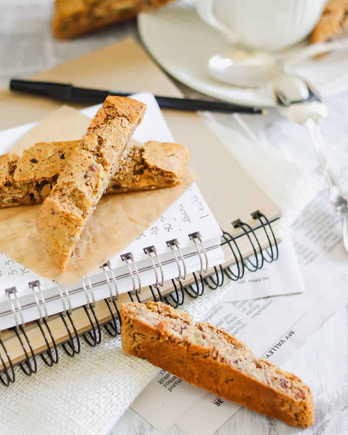 A notebook, pen, and almond biscotti are arranged on a table alongside a coffee cup and spoon.