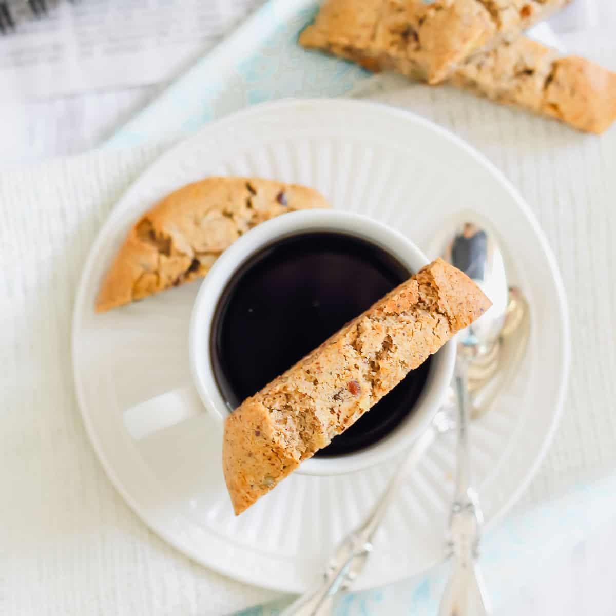 A cup of black coffee with almond biscotti on a white saucer, accompanied by two silver spoons. More biscotti pieces are in the background.