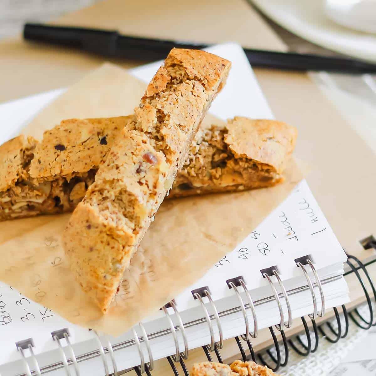 Two biscotti cookies rest on parchment paper placed on an open spiral notebook, with a pen in the background.