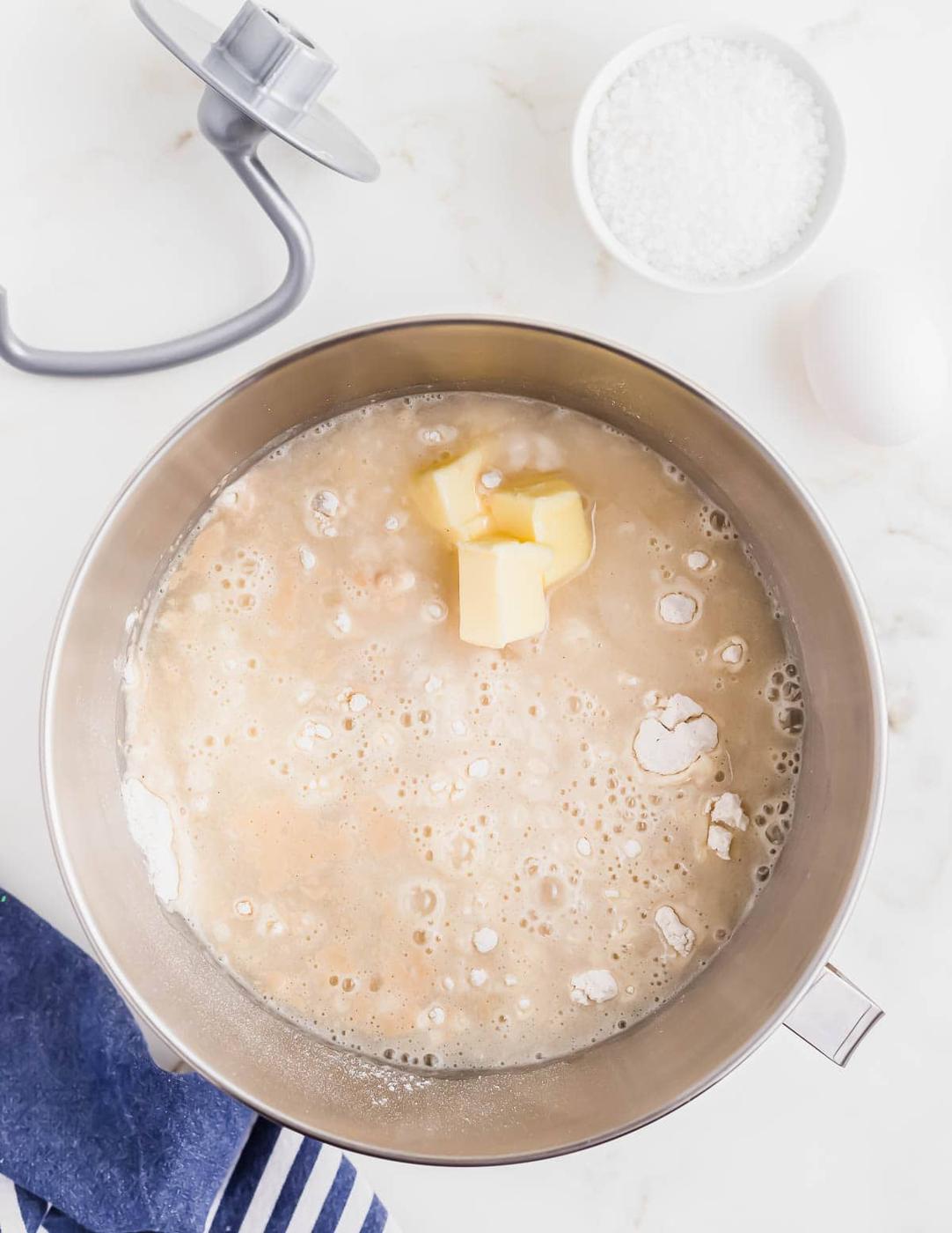 A mixing bowl with flour, water, butter, and other ingredients. Nearby are a mixer attachment, a small bowl of coarse salt, an egg, and a blue cloth on a white countertop.