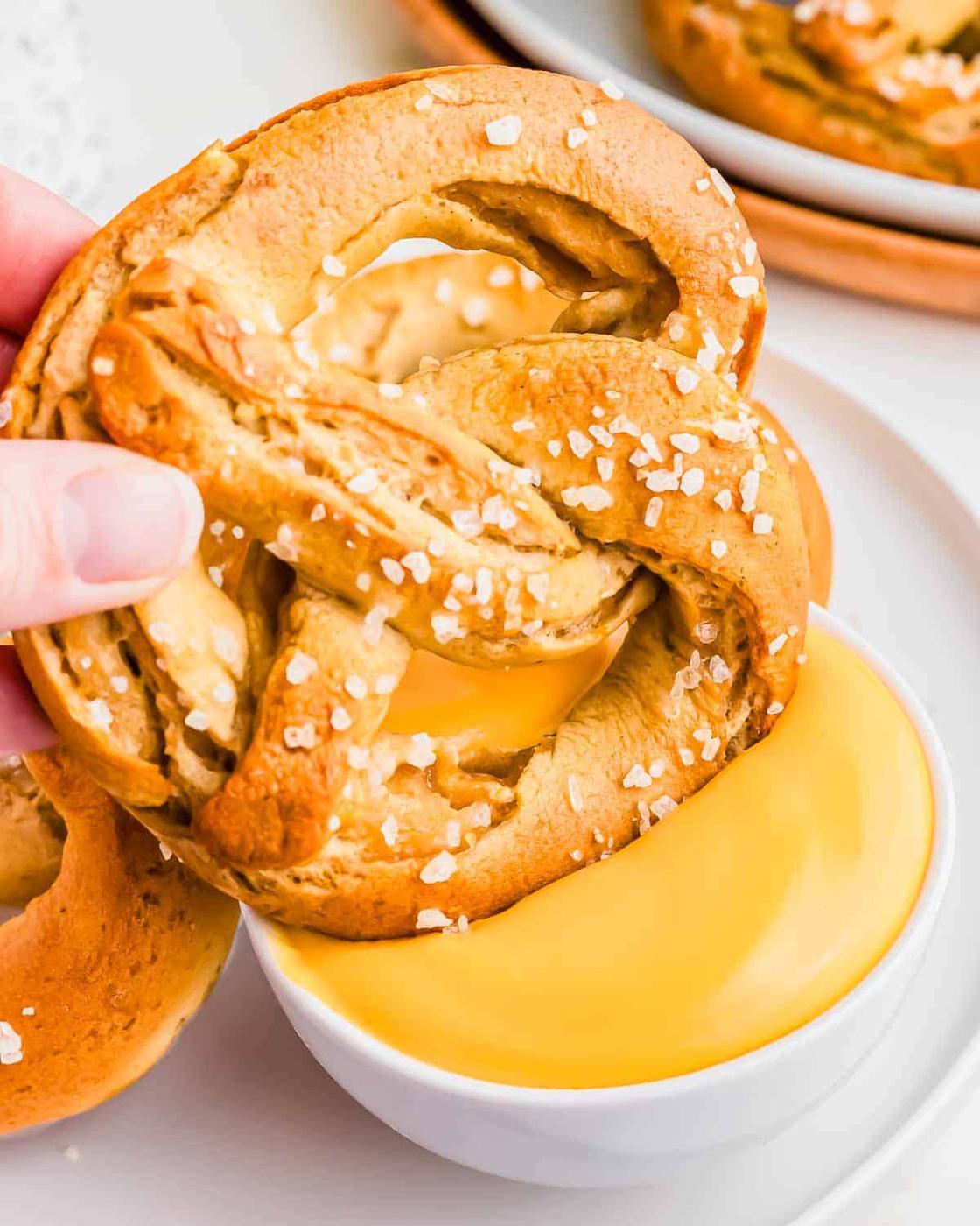 A hand holding a pretzel over a bowl of cheese sauce.