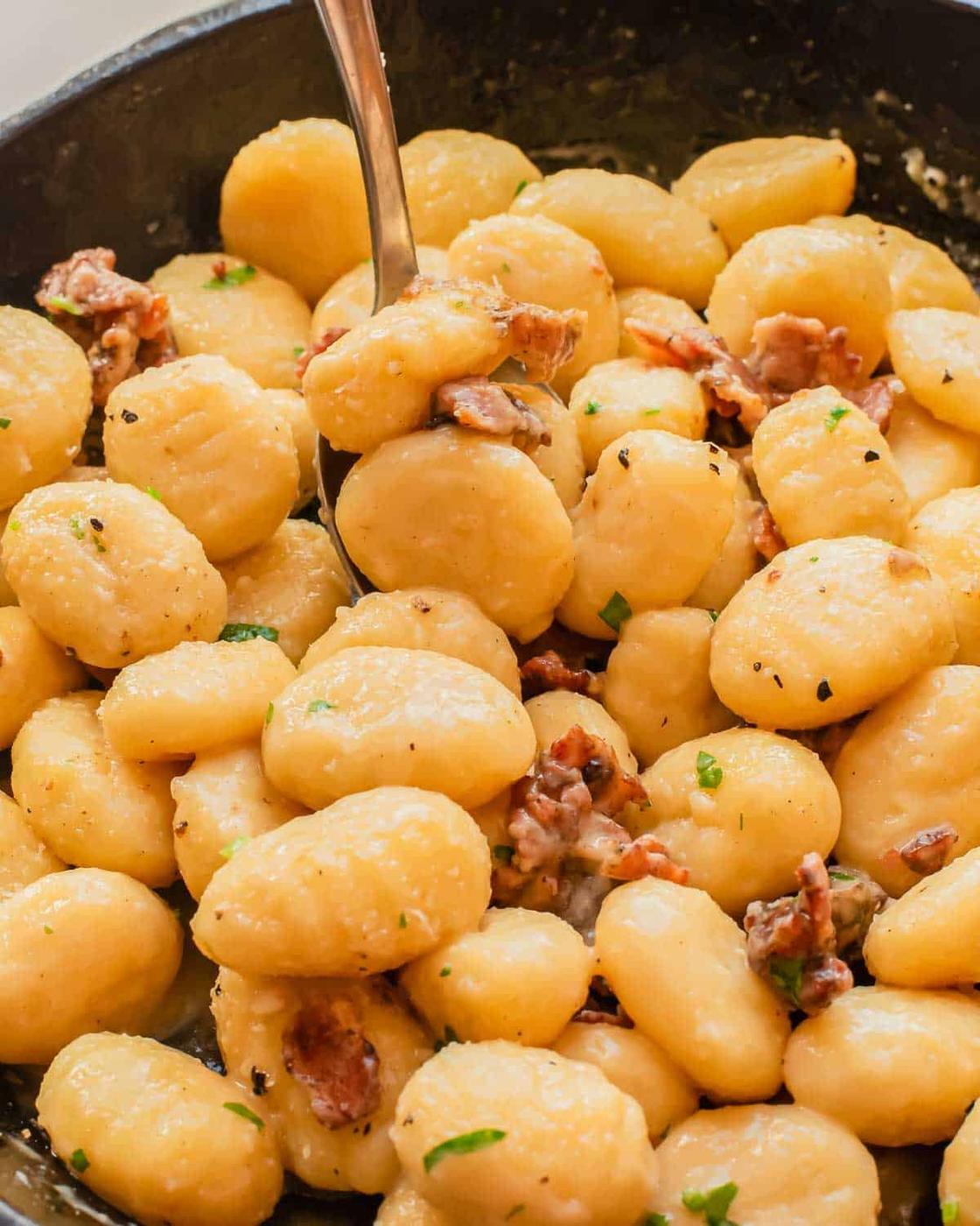A close-up of carbonara gnocchi mixed with small pieces of crispy bacon and garnished with chopped herbs, served in a black skillet with a spoon.