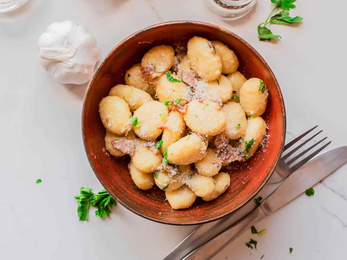A bowl of gnocchi topped with grated cheese and herbs sits on a white table, surrounded by garlic, chopped parsley, a fork, and a knife.