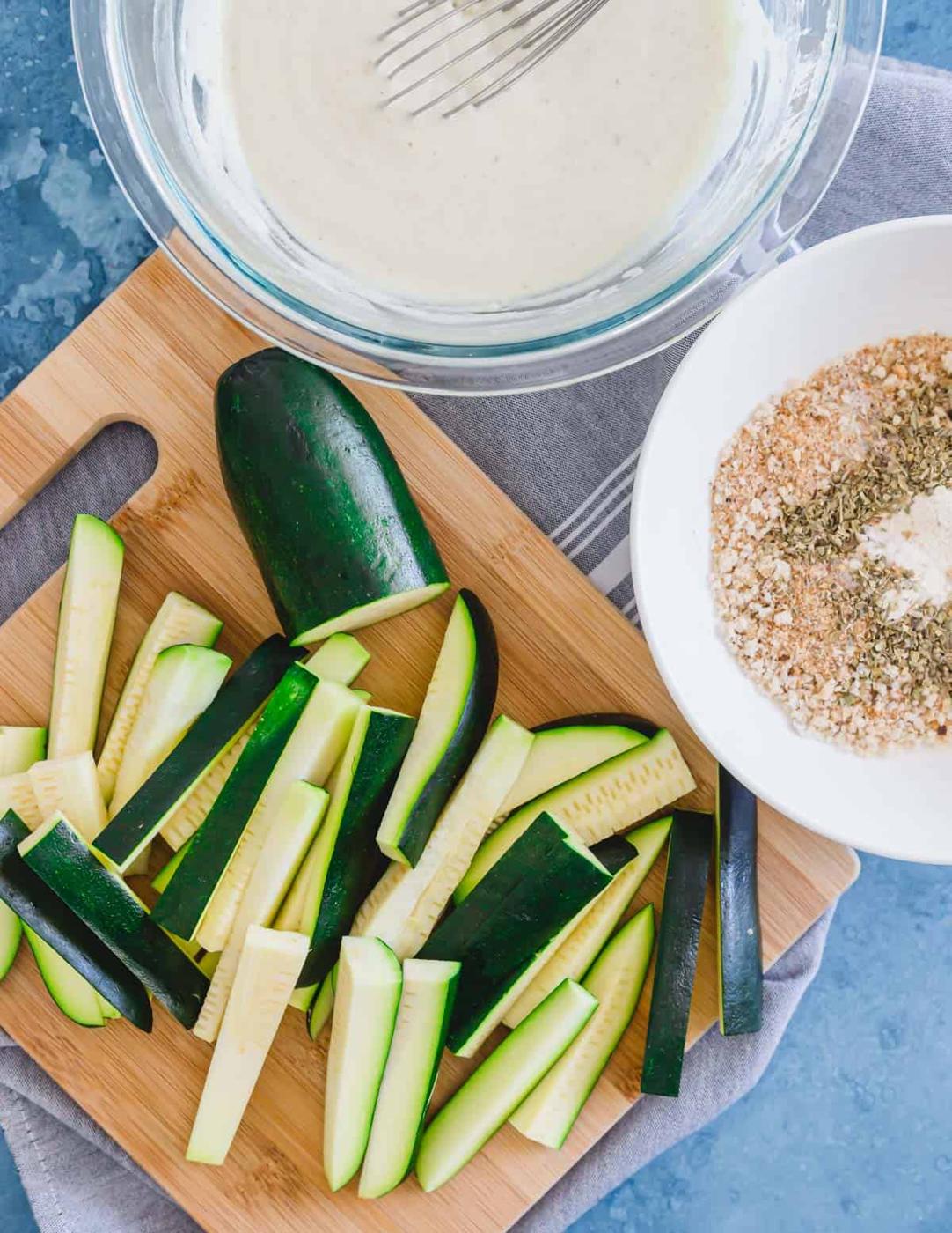 Sliced zucchini on a cutting board with breadcrumb mixture and batter in bowls on the side.