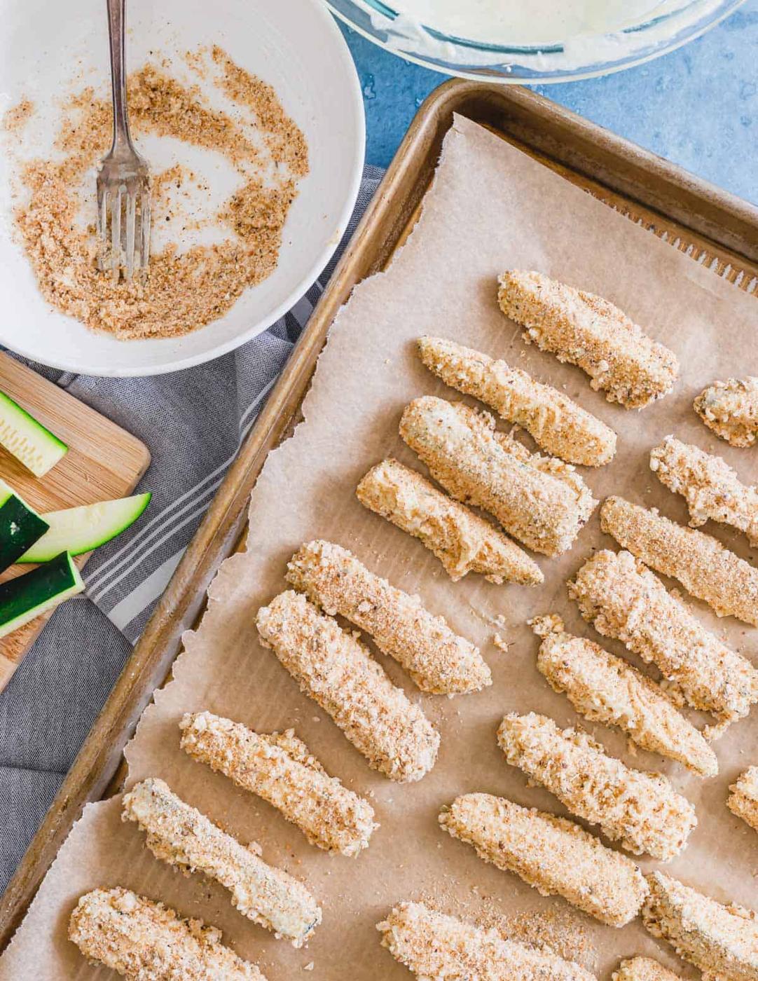 Battered zucchini fries lined up on a parchment covered baking sheet.