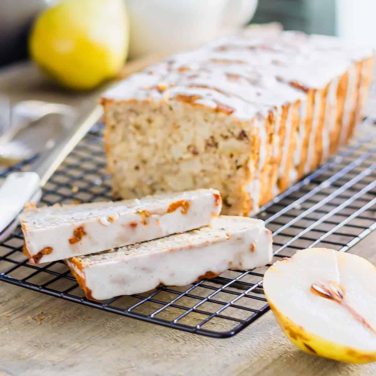 A loaf of glazed pear bread with two slices cut, placed on a cooling rack next to half a pear.
