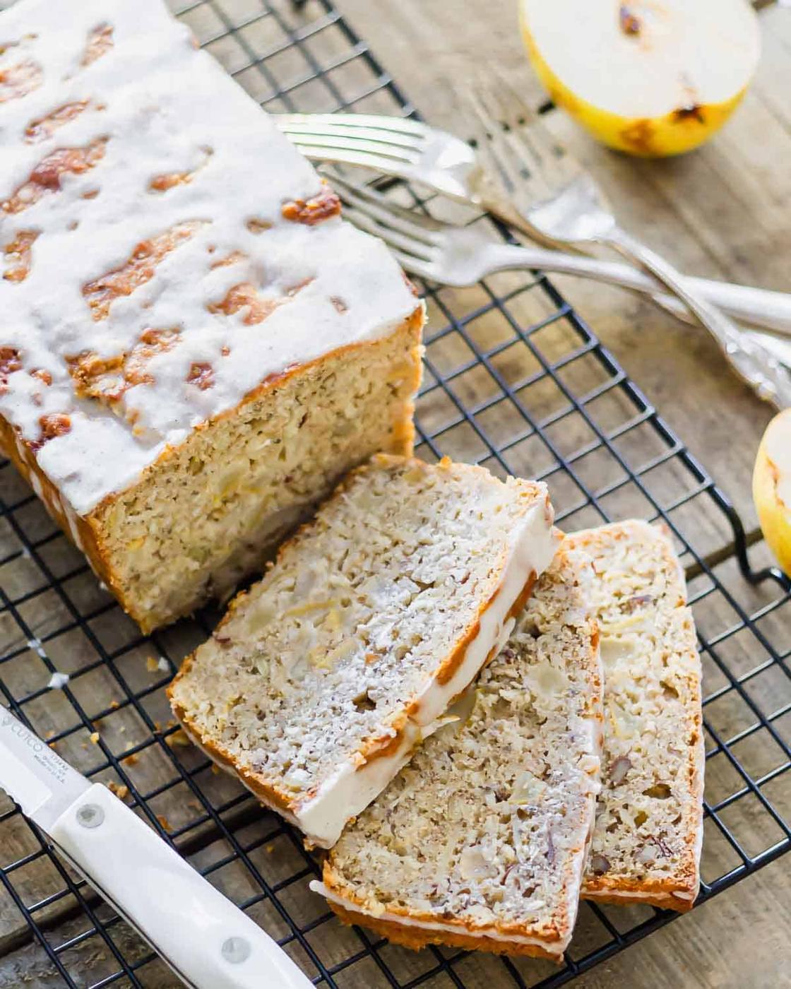 A loaf of granola pear bread with white glaze sits on a cooling rack. Two slices have been cut, and a knife lies nearby. A halved pear is visible in the background. Two forks are also present.