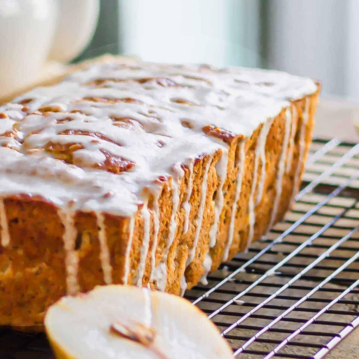 A loaf of bread with white icing drizzle on a cooling rack. A halved pear is visible in the foreground.