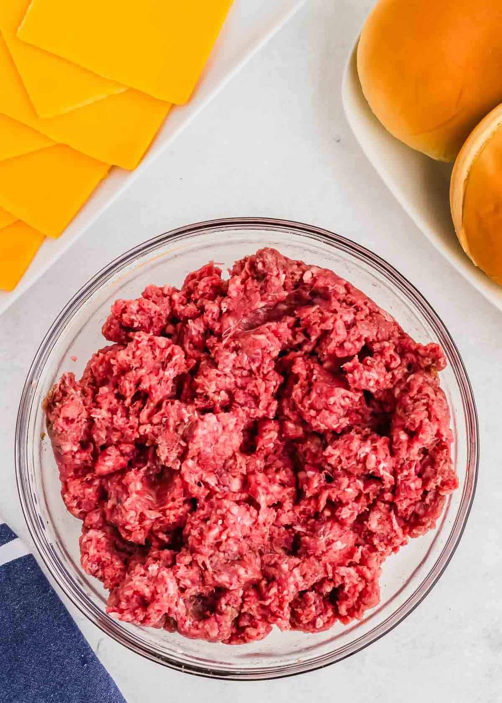 A bowl of raw ground bison on a kitchen counter, accompanied by slices of cheese and burger buns.