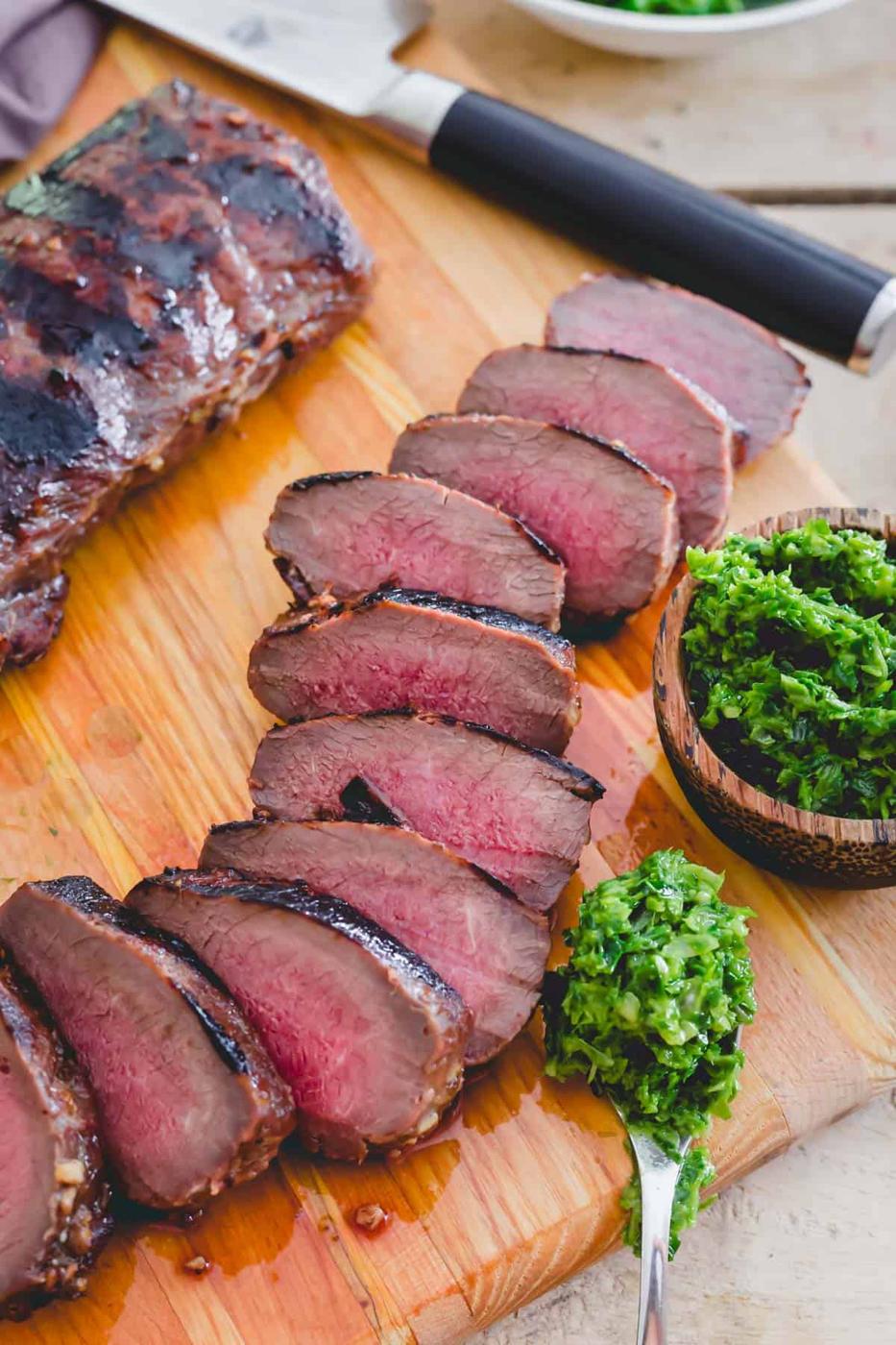Sliced medium-rare venison backstrap, expertly grilled with a dark char on the edges, rests on a wooden cutting board. A small wooden bowl brimming with chopped green herbs sits nearby, accompanied by a spoon holding more herbs—a perfect recipe for flavor.