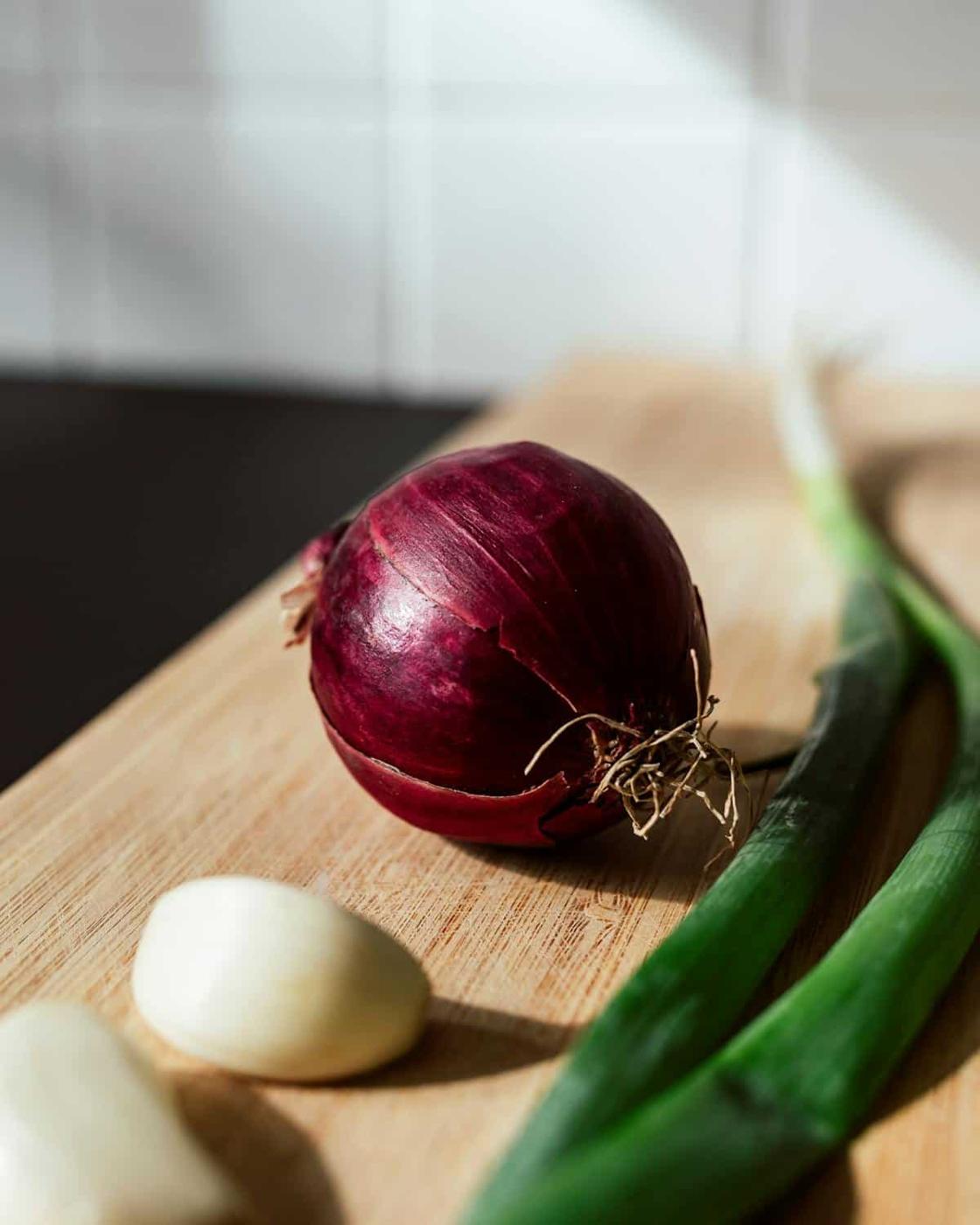 A red onion, a whole garlic clove, and a green onion on a wooden cutting board, with a tiled wall in the background.