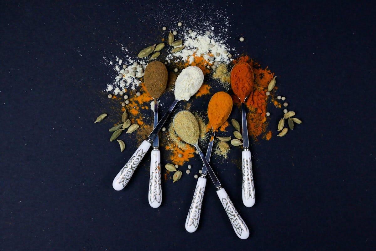 Five ornate spoons with white handles each hold different spices—brown, white, orange, and red—on a dark surface. Spices and cardamom pods are artistically scattered around the spoons.