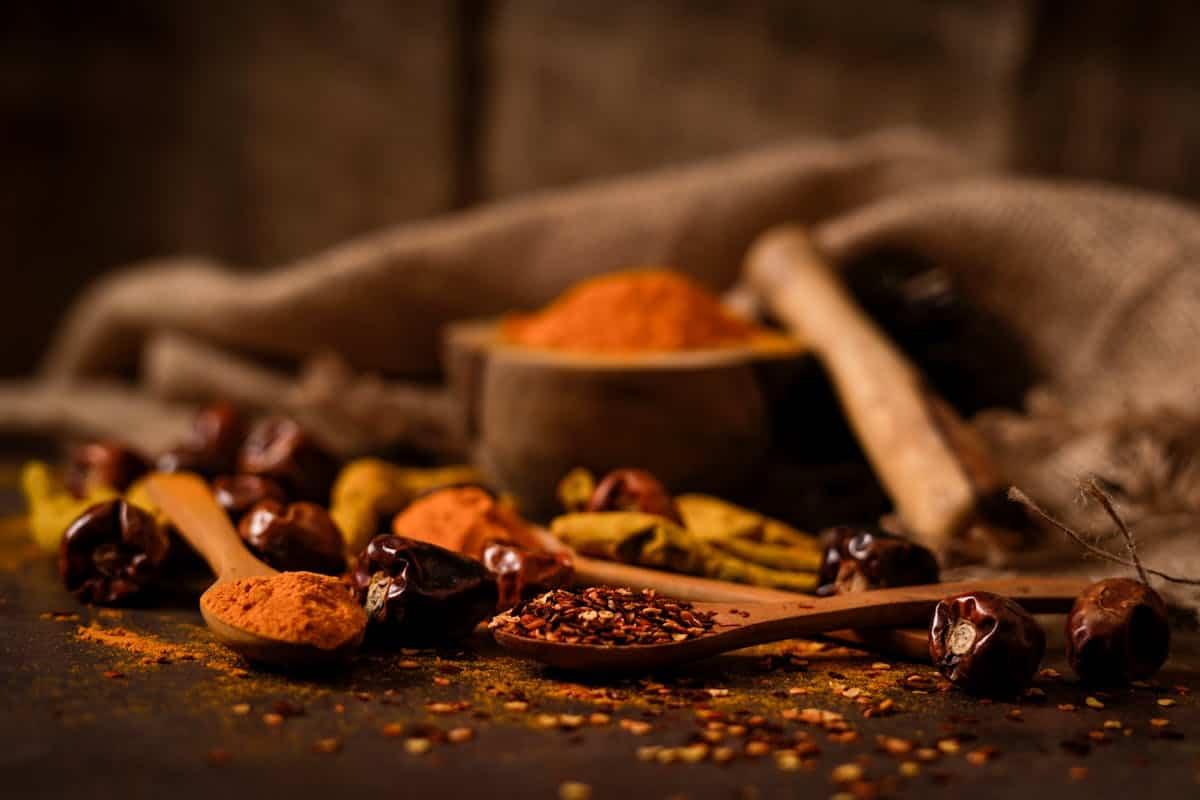 Wooden spoons holding orange and brown spices are surrounded by whole dried chilies and seeds. A small wooden bowl filled with turmeric is in the background, with a rustic cloth draped nearby on a dark, textured surface.