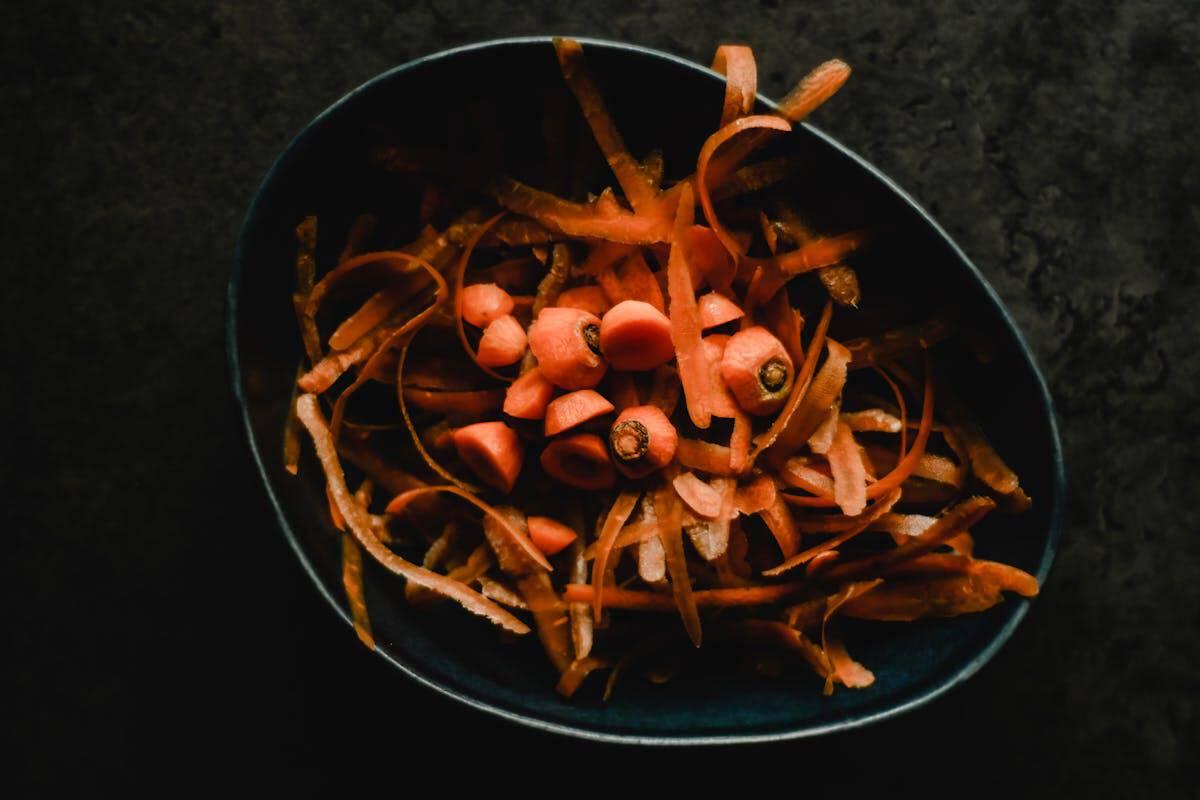 A dark bowl filled with spirals and chunks of orange carrot peels, nestled in a rich homemade vegetable broth, set against a dark background.