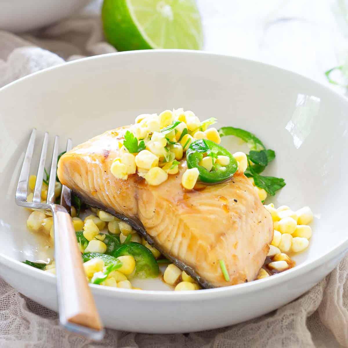A plate with a cooked sablefish fillet topped with corn and jalapeno slices, garnished with herbs. A fork rests beside the salmon, and a half lime is in the background.