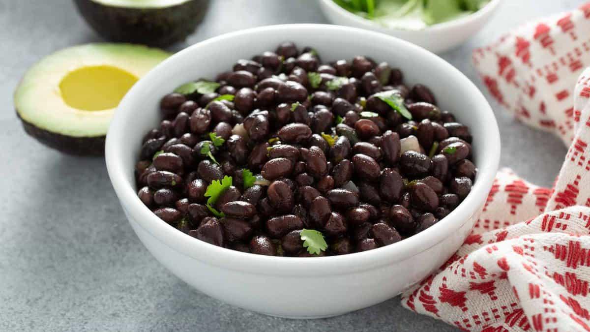A white bowl filled with cooked black beans garnished with cilantro, placed on a grey surface next to half an avocado and a red-and-white cloth.