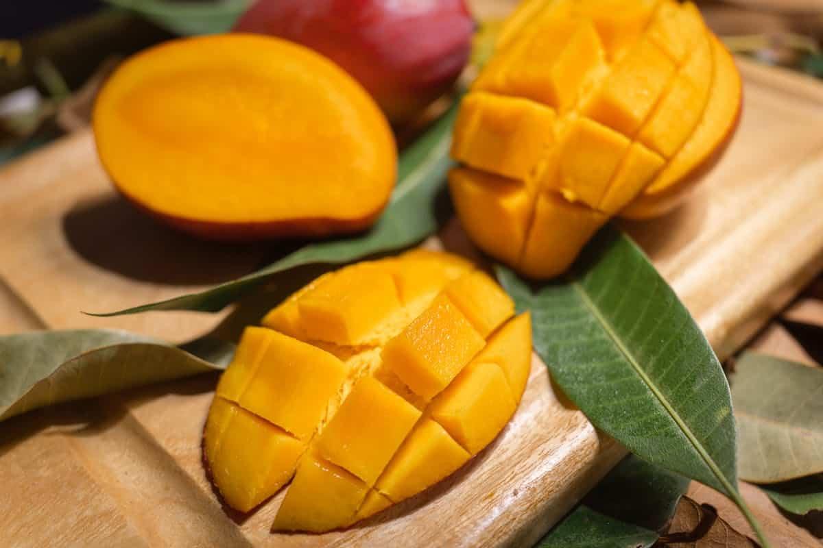 Learn how to cut a mango with ease: Sliced mango pieces and a half mango displayed on a wooden board with green leaves.