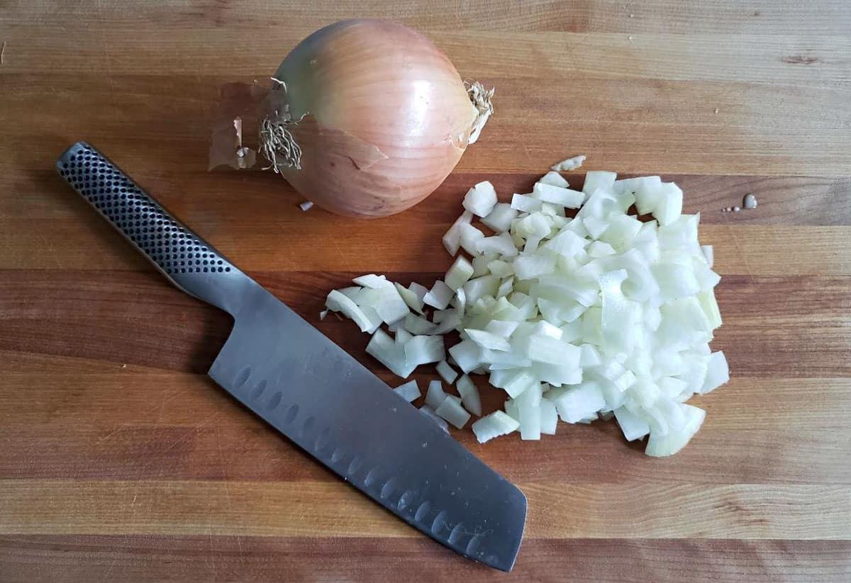 Whole yellow onion next to diced onion with a knife on a cutting board.