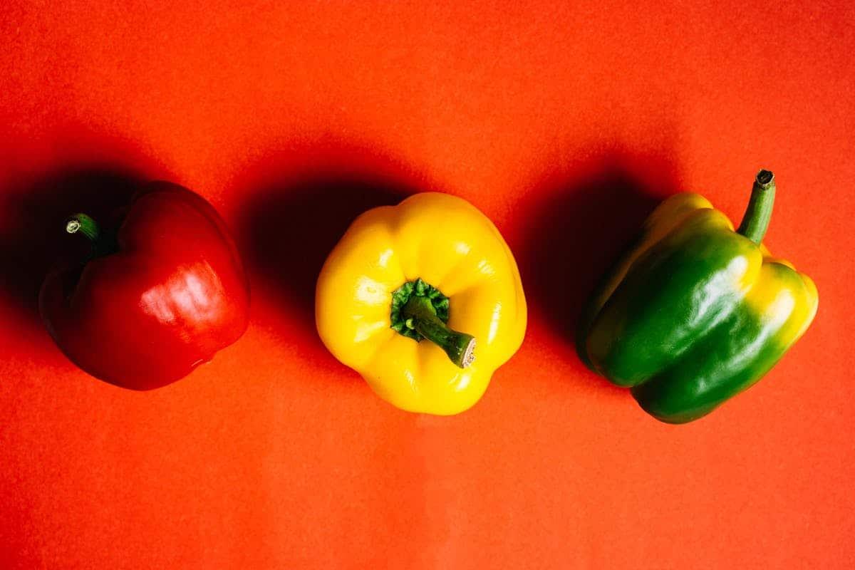 A red bell pepper, a yellow bell pepper, and a green bell pepper are lined up horizontally on a bright orange background, offering inspiration for learning how to freeze peppers effectively.
