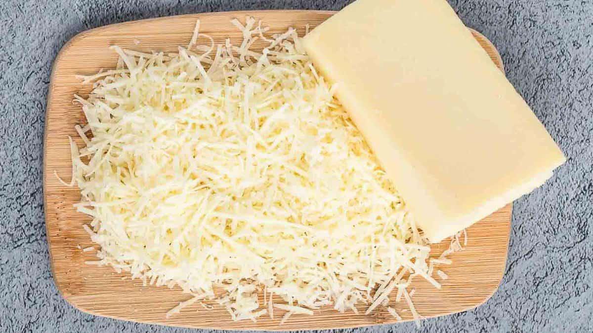 A wooden board with a block of cheese and a pile of shredded cheese on a textured gray surface.
