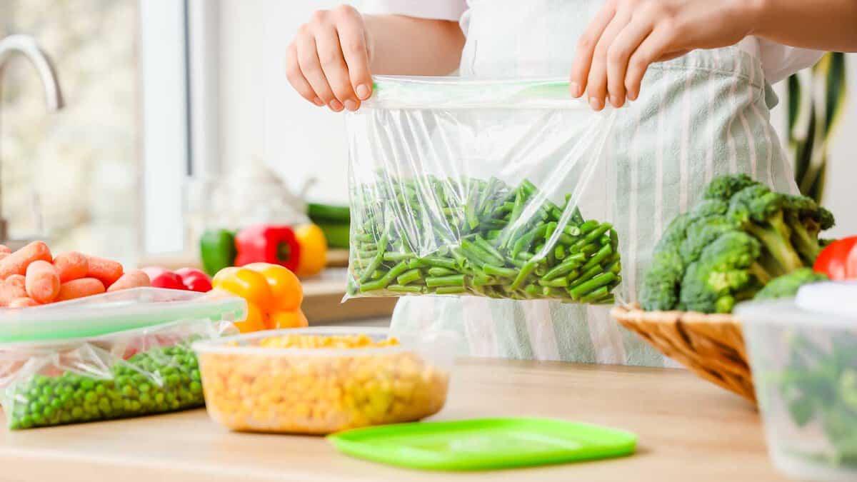 A person in a kitchen holds a clear plastic bag of green beans. The countertop displays various vegetables, including carrots, peas, corn, yellow peppers, and broccoli. The scene is bright and well-organized, suggesting meal prep.