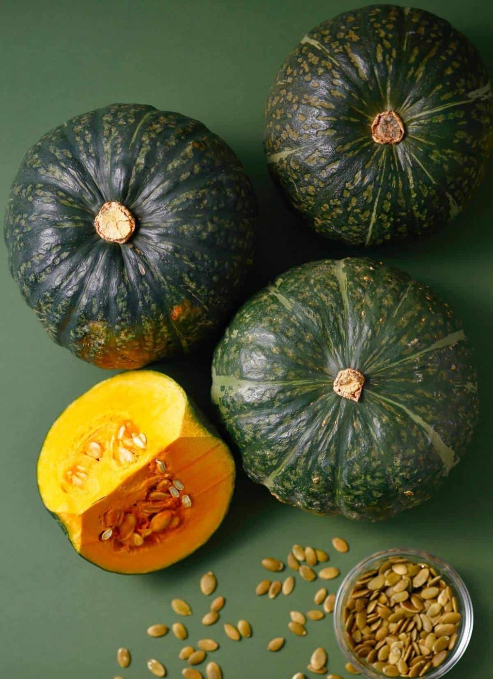 Three whole green kabocha squashes, one cut open showing its yellow-orange flesh and seeds, with a small glass bowl of pumpkin seeds scattered nearby on a green background.