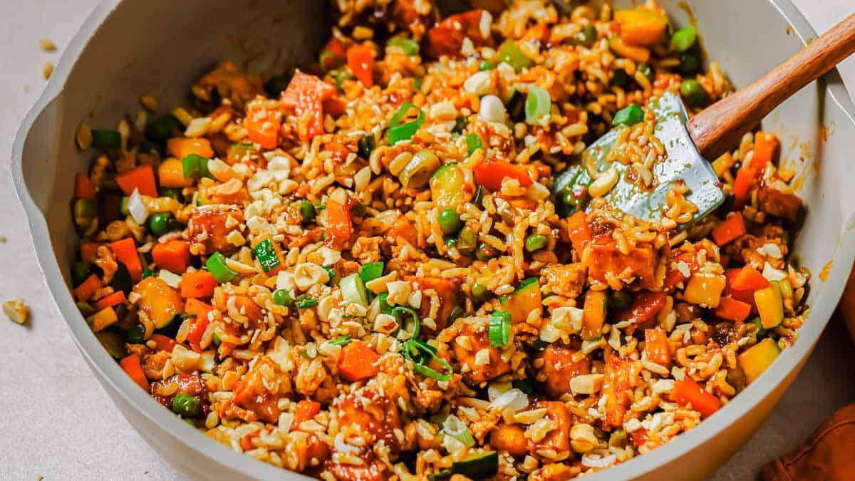 A pan full of tofu fried rice and vegetables with a wooden spoon.