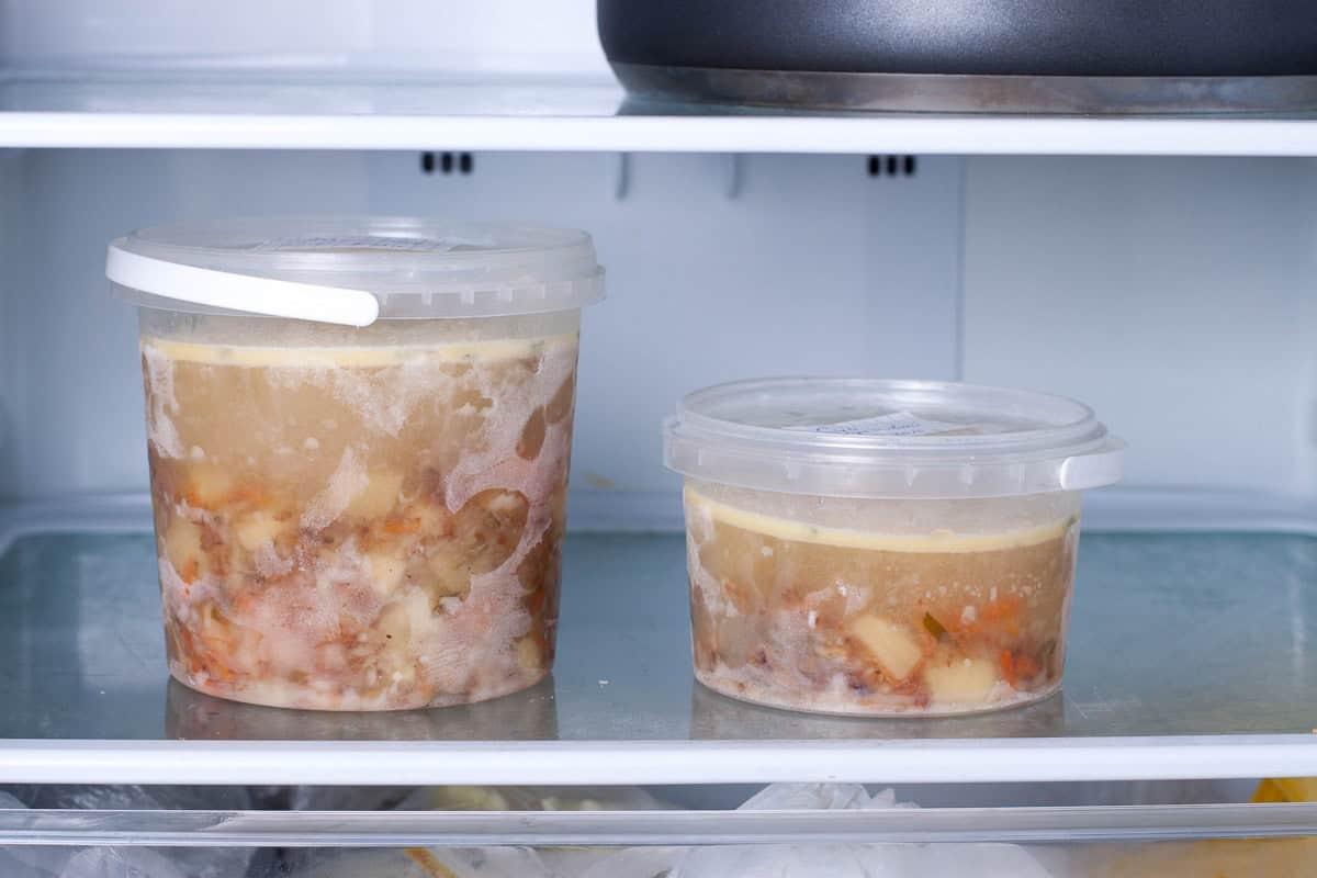 Two plastic containers with lids hold broth with chunks of vegetables and meat, sitting on a refrigerator shelf. The contents are slightly congealed from refrigeration. A black pot is partially visible on the shelf above.