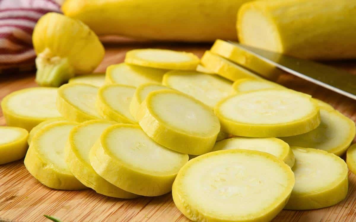 Sliced yellow squash on a wooden cutting board, with a knife and whole yellow squash in the background. Learn how to freeze summer squash to preserve its fresh flavor for longer.