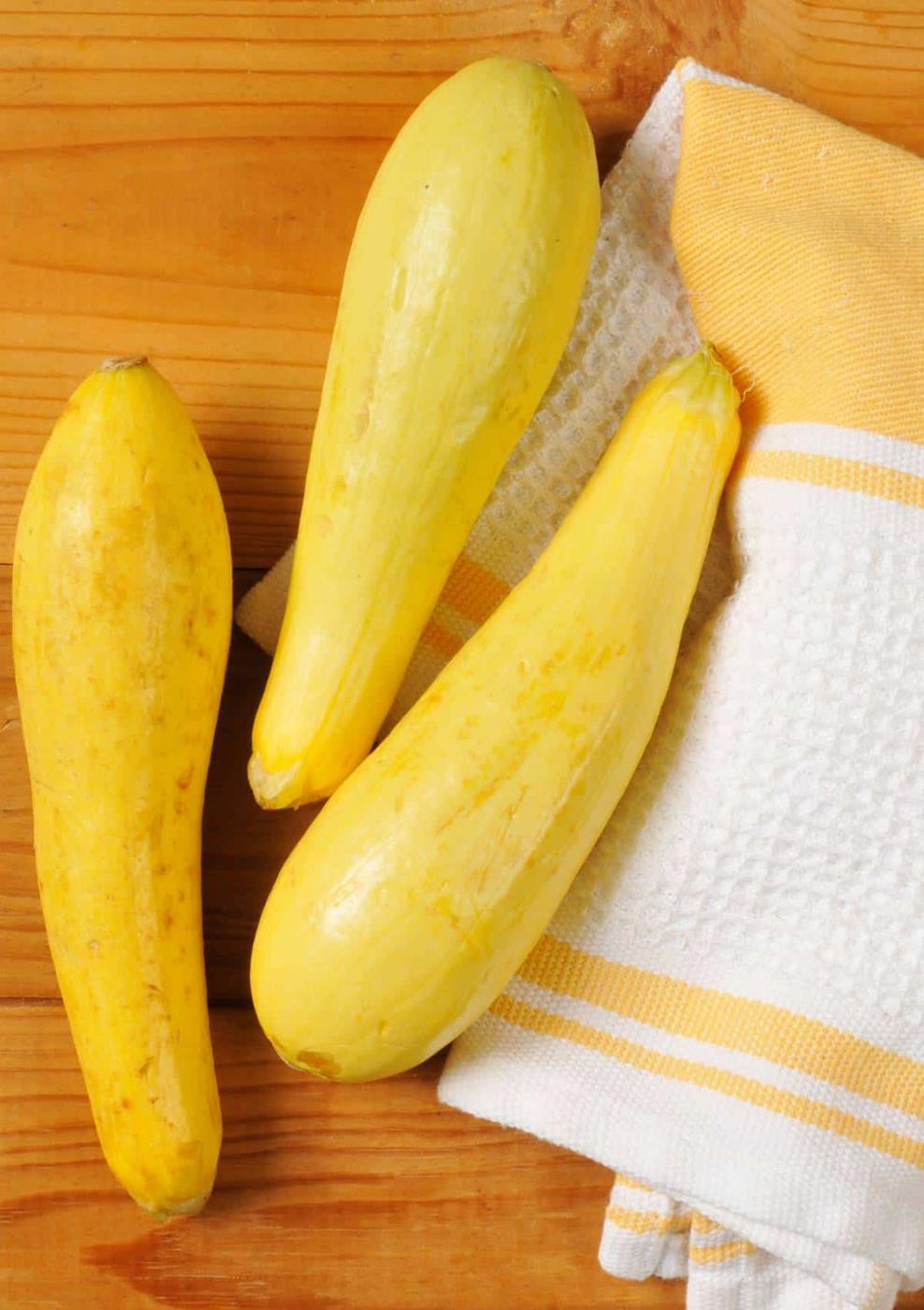 Three yellow squash lie on a wooden surface next to a white and yellow striped kitchen towel, perfect props for learning how to freeze summer squash.