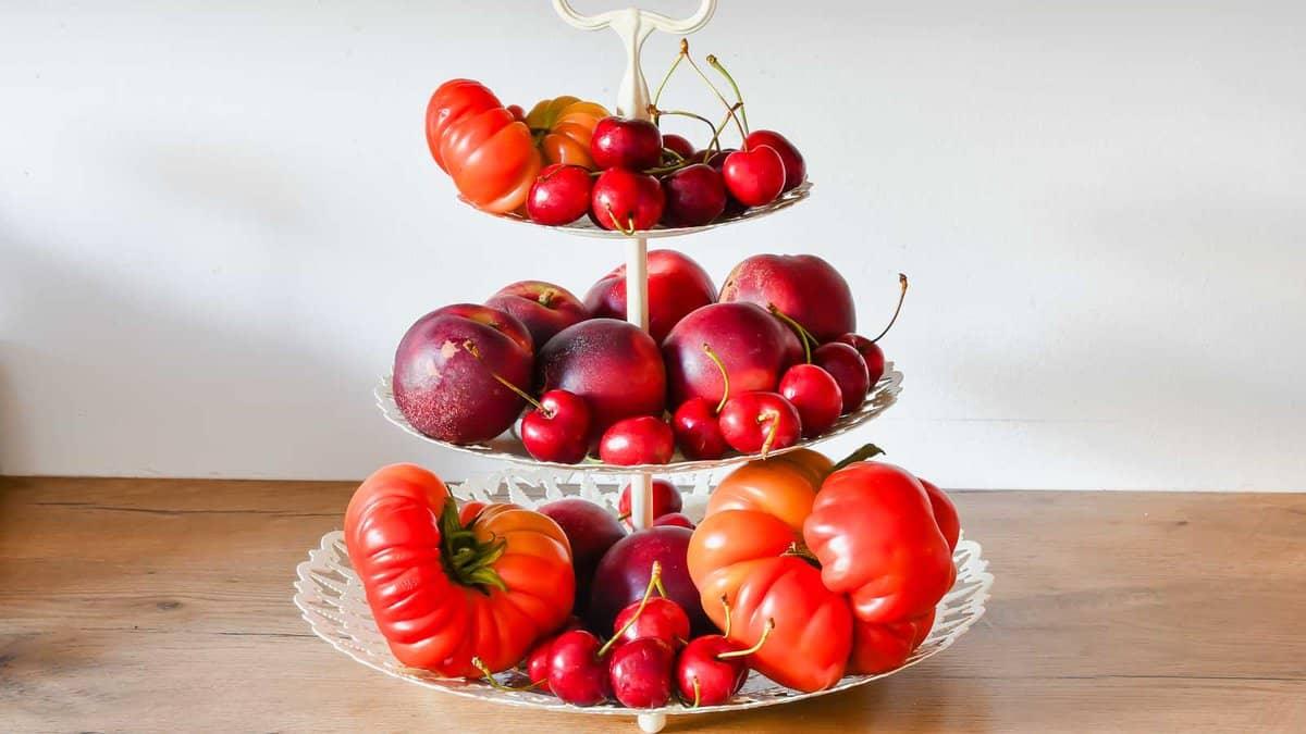 A three-tiered serving tray holds various red fruits, including apples, cherries, and peppers, placed on a wooden surface.