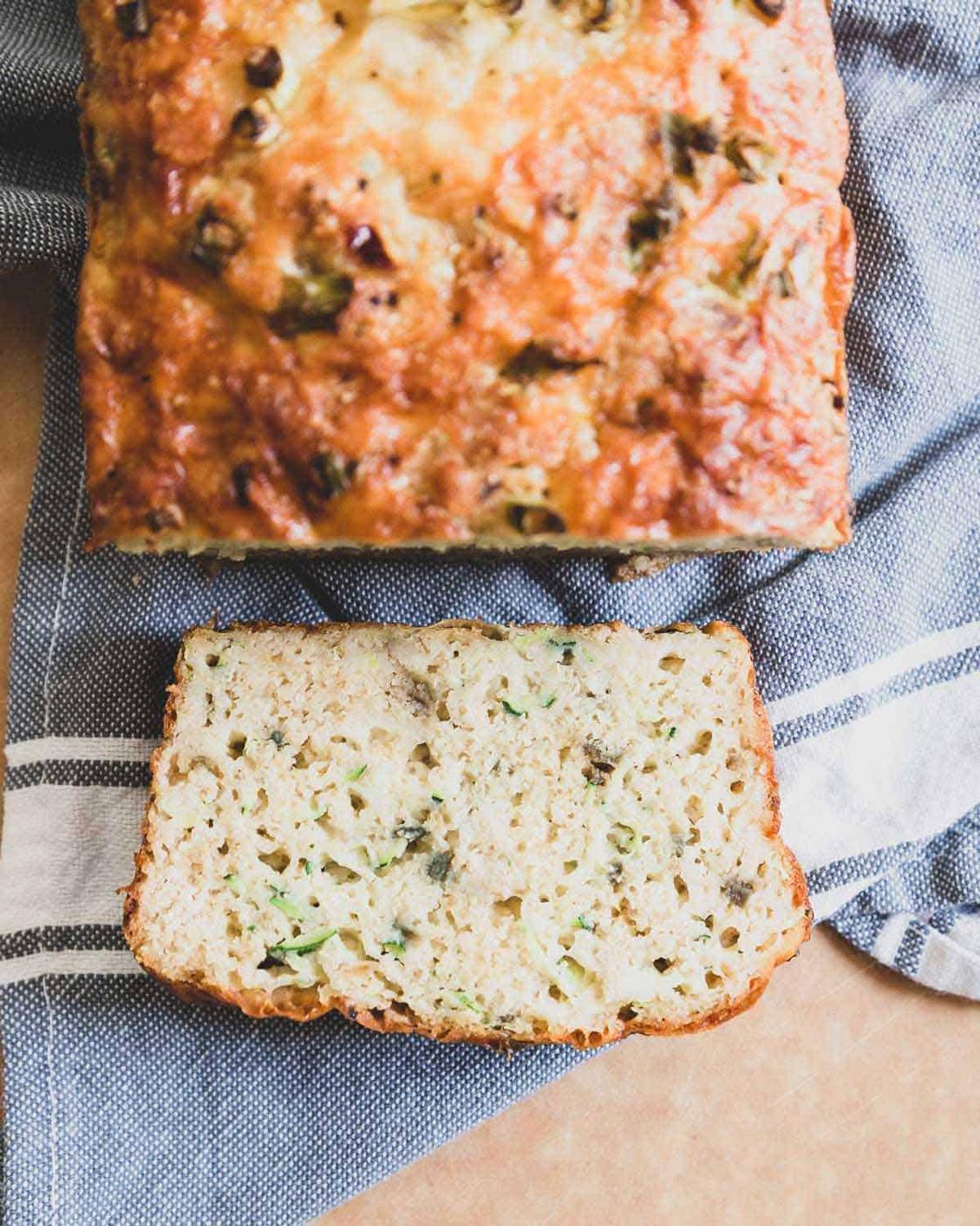 Cheesy zucchini bread on a kitchen towel with one slice laying flat.