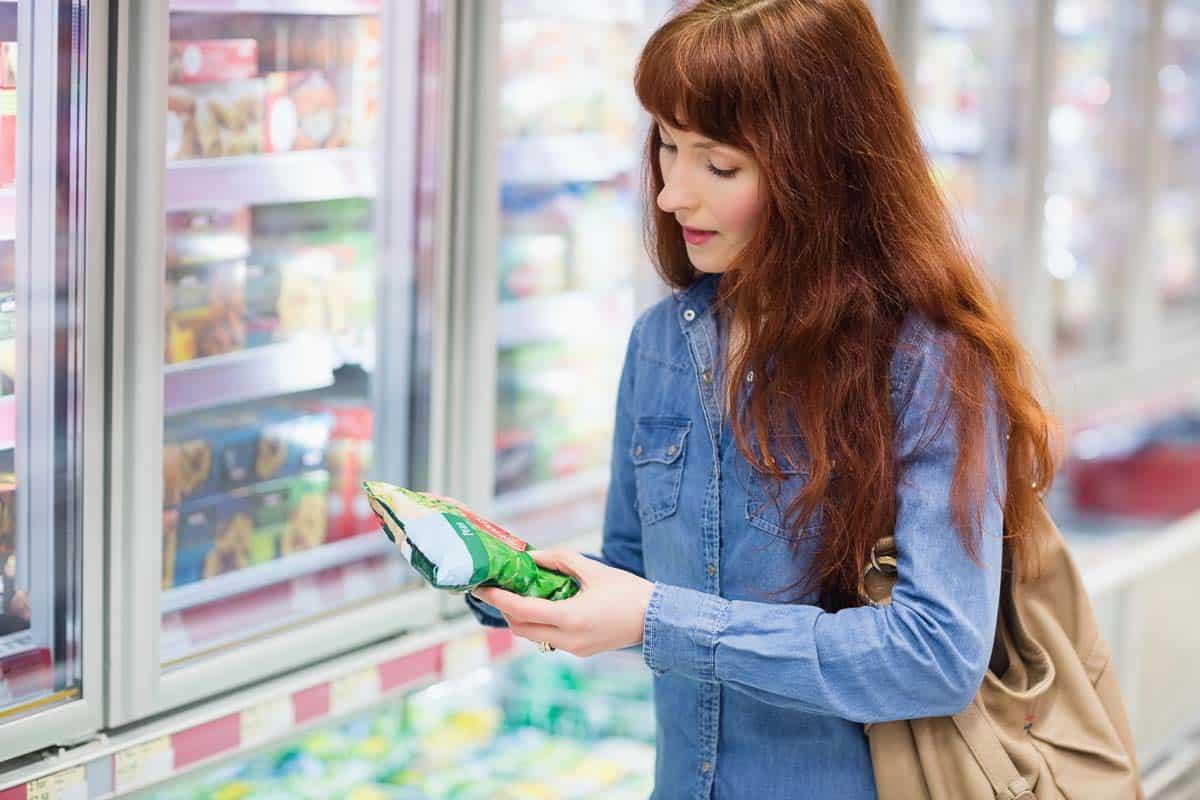 A woman with long red hair stands in a grocery store aisle, holding a bag of frozen vegetables and looking at the packaging. She is wearing a denim shirt and carrying a beige bag.