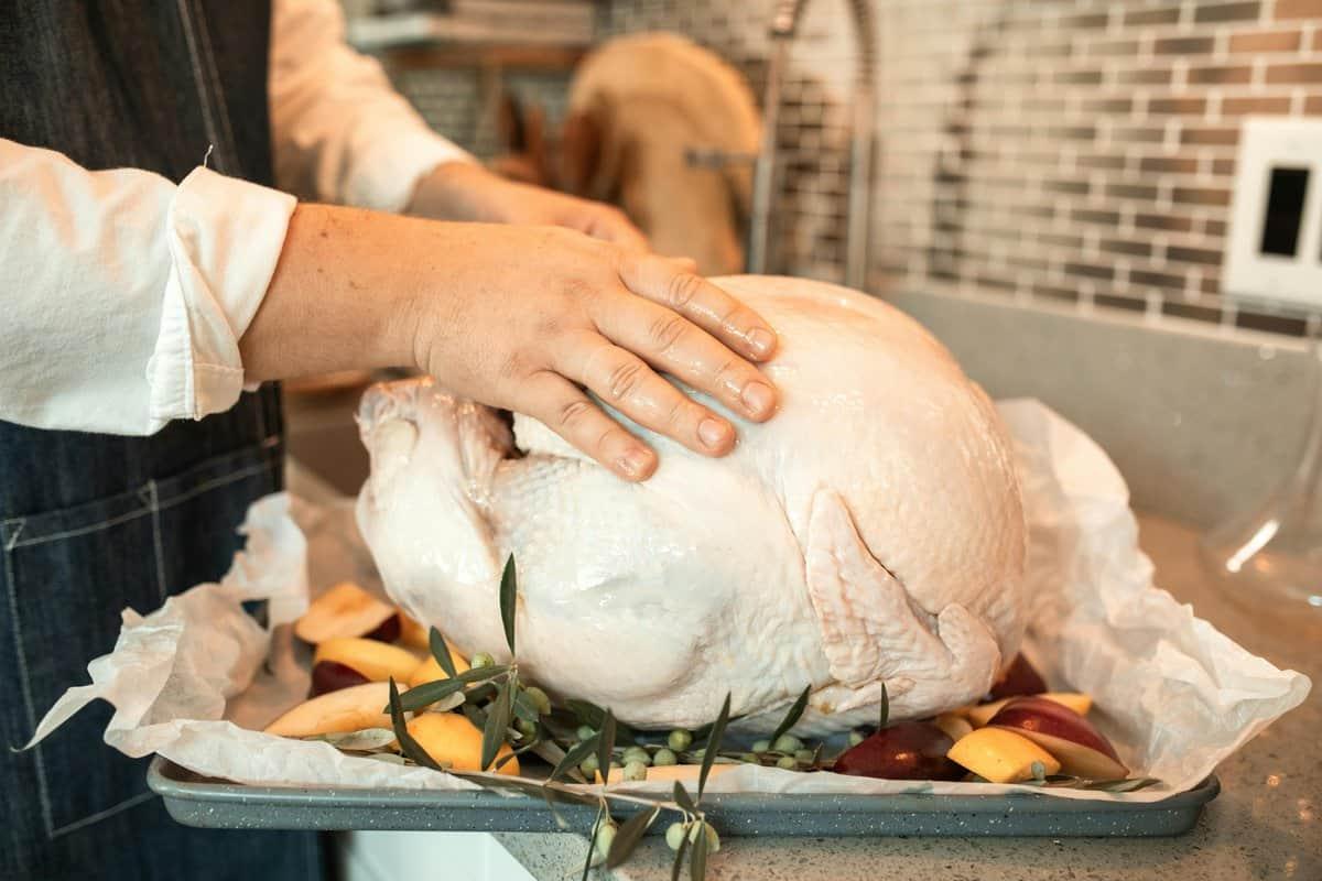 A person prepares a large uncooked turkey on a baking tray, surrounded by sliced apples and herbs, showcasing an essential step before it's ready for roasting—knowing how to thaw a turkey. The kitchen's tiled wall and sink serve as the perfect backdrop for this culinary scene.
