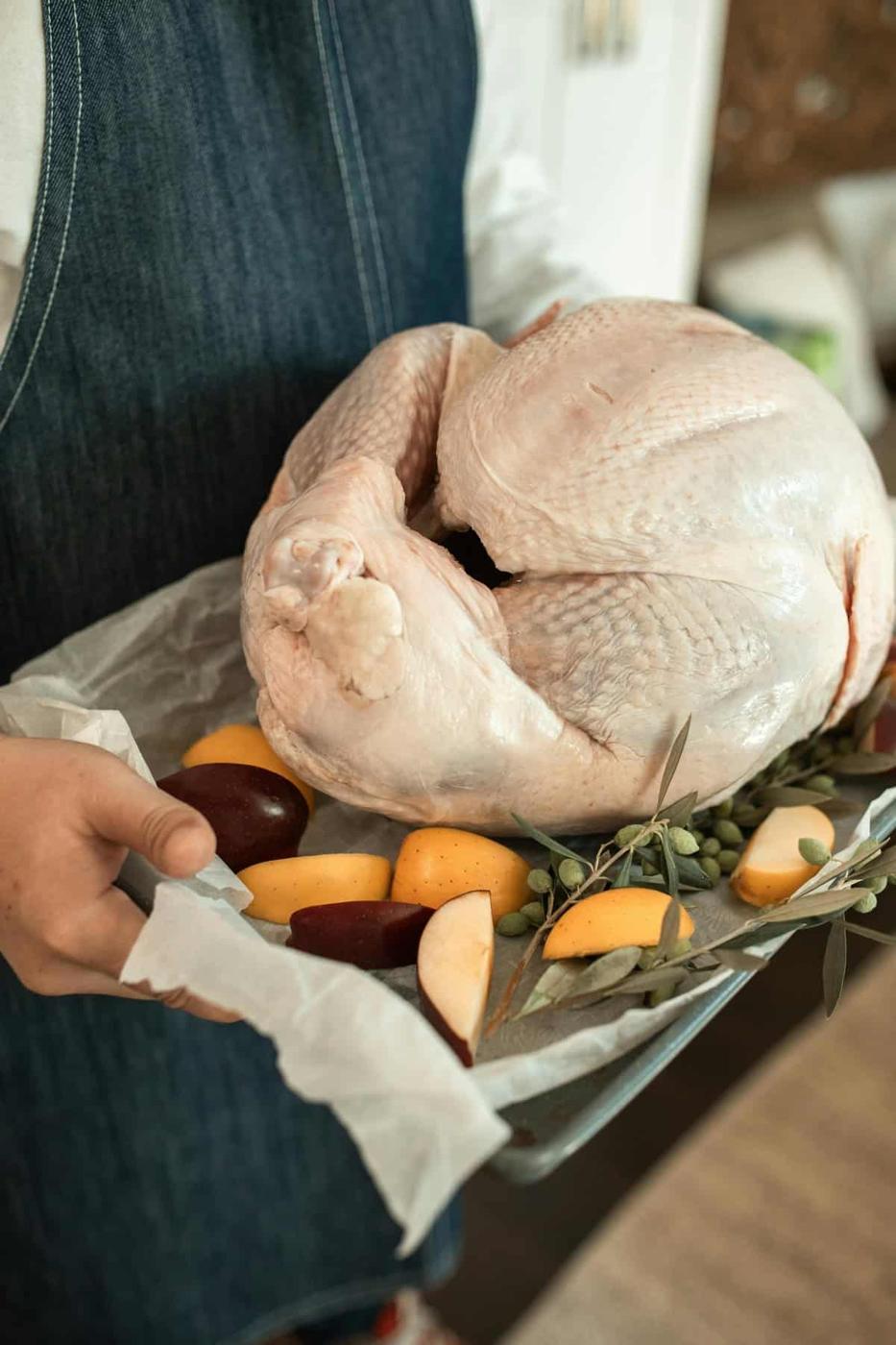 A person in a denim apron holds a baking tray with a raw, well-thawed whole turkey. The tray also contains apple and pear slices, herbs, and small green fruits or olives, ready for cooking.