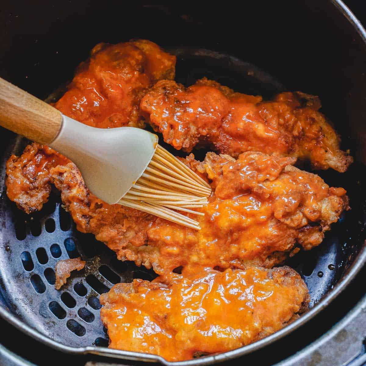 Brushing buffalo sauce onto air fried chicken tenders in the air fryer basket.