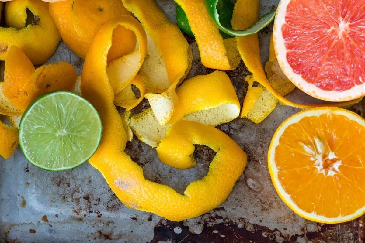 Slices and peels of citrus fruits—lime, orange, lemon, and grapefruit—are spread across a rustic metal surface. The orange and grapefruit are halved, and the yellow peels are curled and scattered.