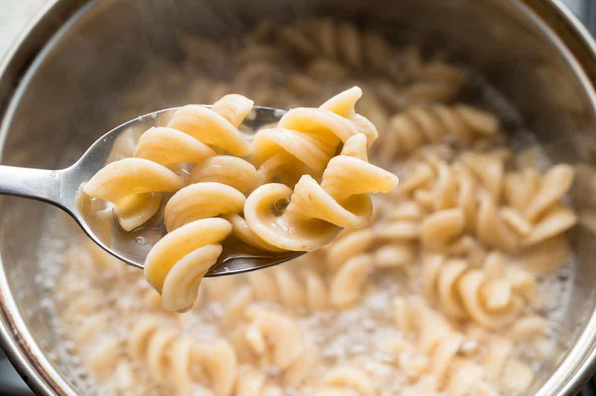 A close-up of a spoon holding cooked rotini pasta above a pot filled with more pasta and boiling water. Steam is rising from the pot.