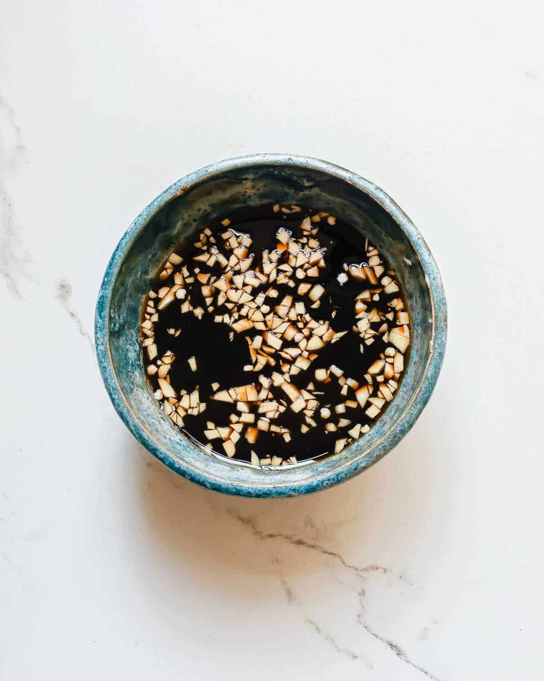 A small blue bowl filled with soy sauce and finely chopped garlic, placed on a white marble surface.