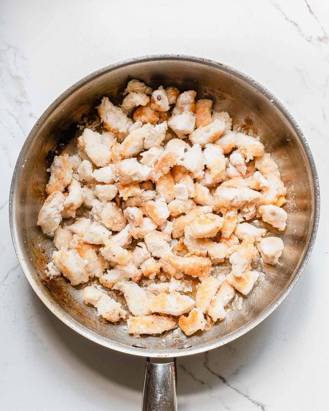 A stainless steel frying pan filled with cooked, browned pieces of chicken on a white countertop.