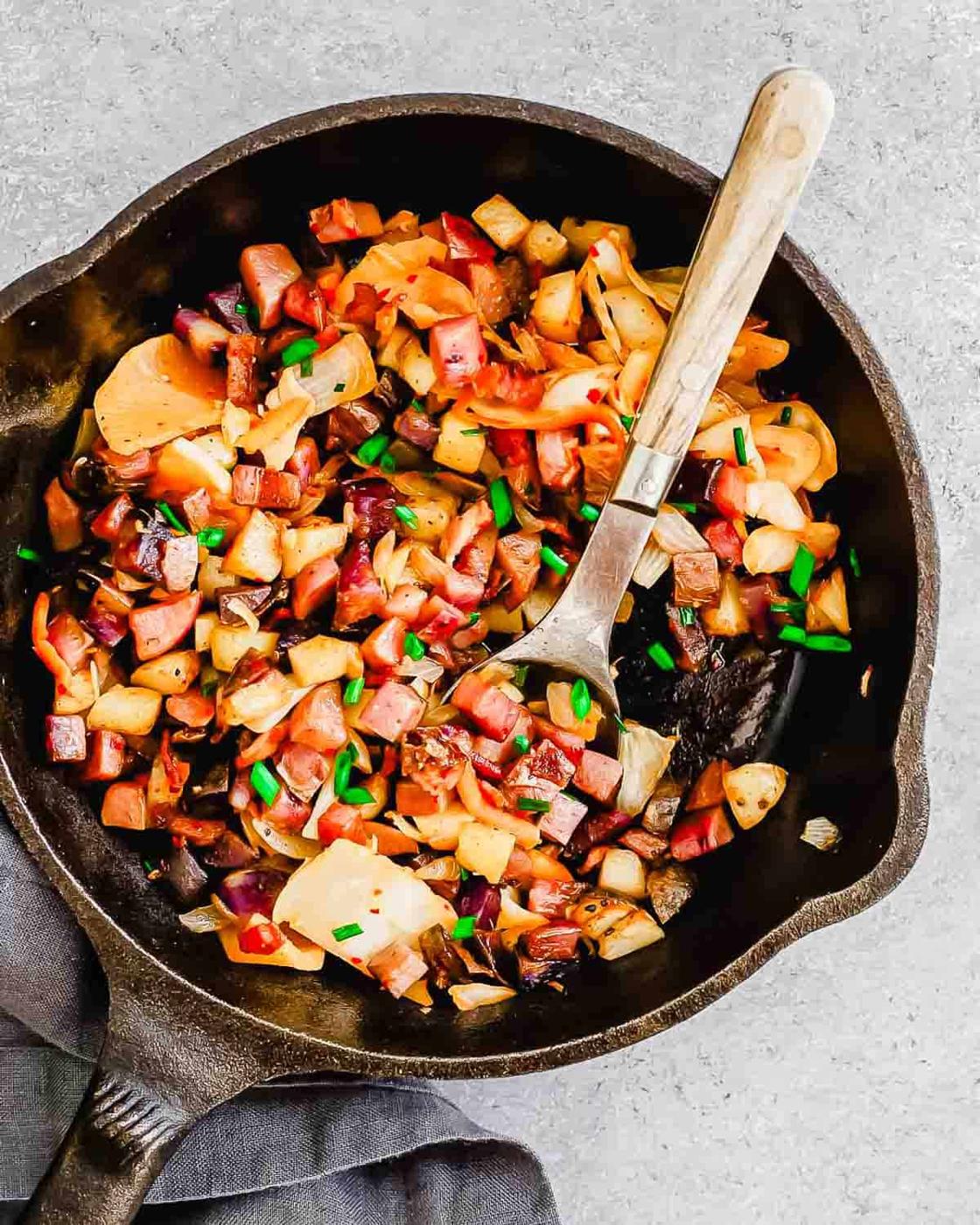 A cast-iron skillet filled with cooked diced potatoes, onions, and pieces of ham, garnished with chopped green onions, is displayed with a silver spoon on the side.