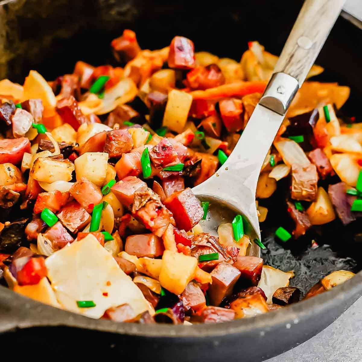 A close-up of a skillet filled with a cooked potato hash dish featuring diced vegetables, meat, and garnished with chopped green herbs. A metal spatula rests in the skillet.