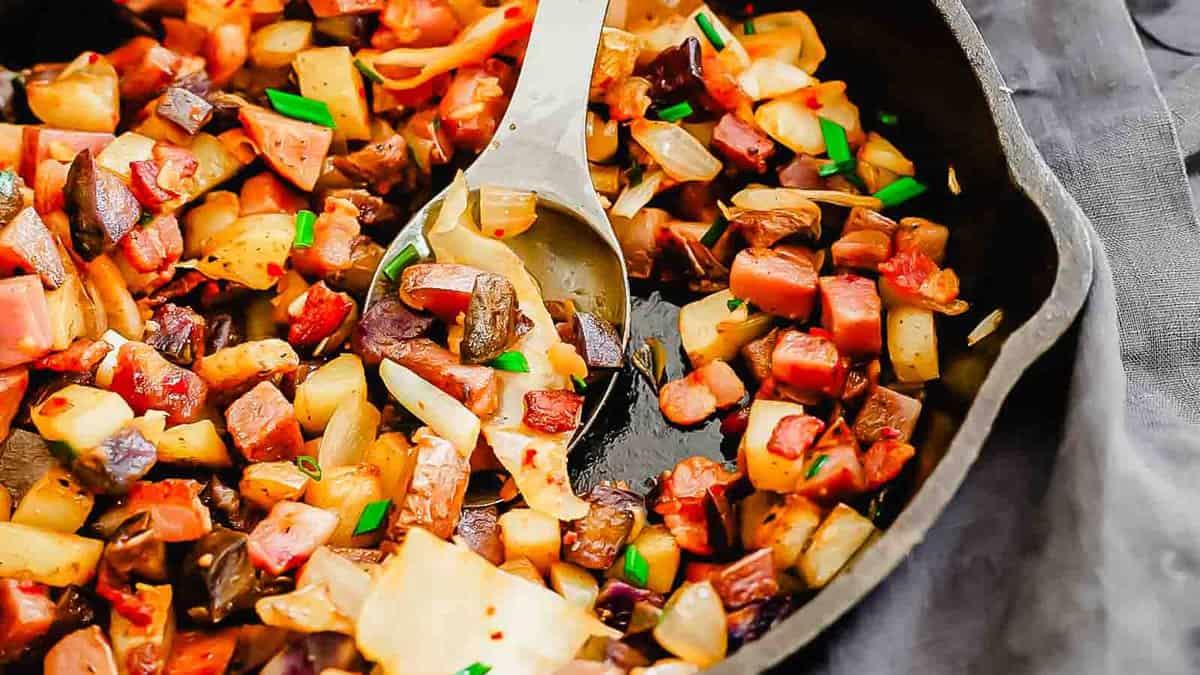 A potato hash skillet filled with diced vegetables and meat, cooked and mixed together, with a serving spoon placed in the center.
