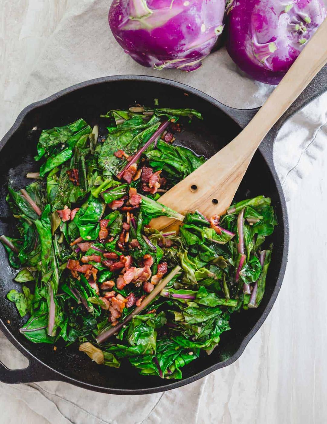 Sautéed kohlrabi greens in a skillet with crispy bacon.