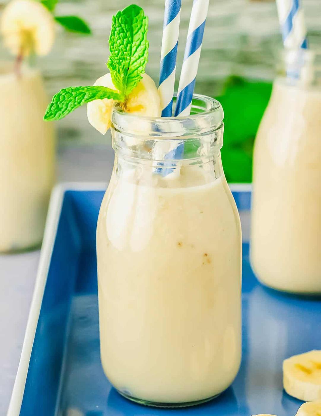 A glass bottle filled with creamy milk, garnished with a banana slice and mint sprig. Two striped straws are in the bottle, which is placed on a blue tray with another smoothie in the background.